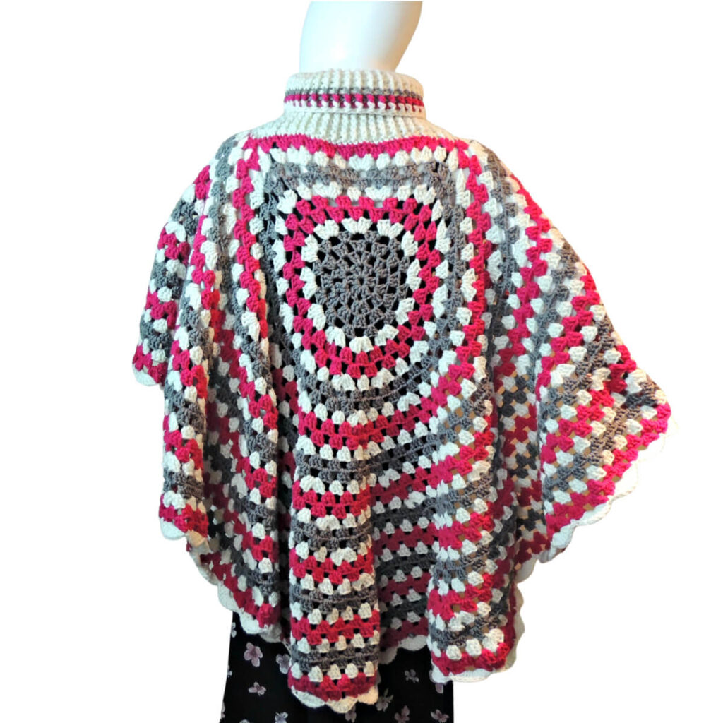 Back view of a mannequin in a black skirt and a poncho made of a large crochet granny stitch circle, the turtleneck of the poncho is about half way from the center to the edge, so that one side of the poncho is much shorter than the other. The poncho is made in stripes of gray, white, and dark pink. The long side of the poncho is in the back.