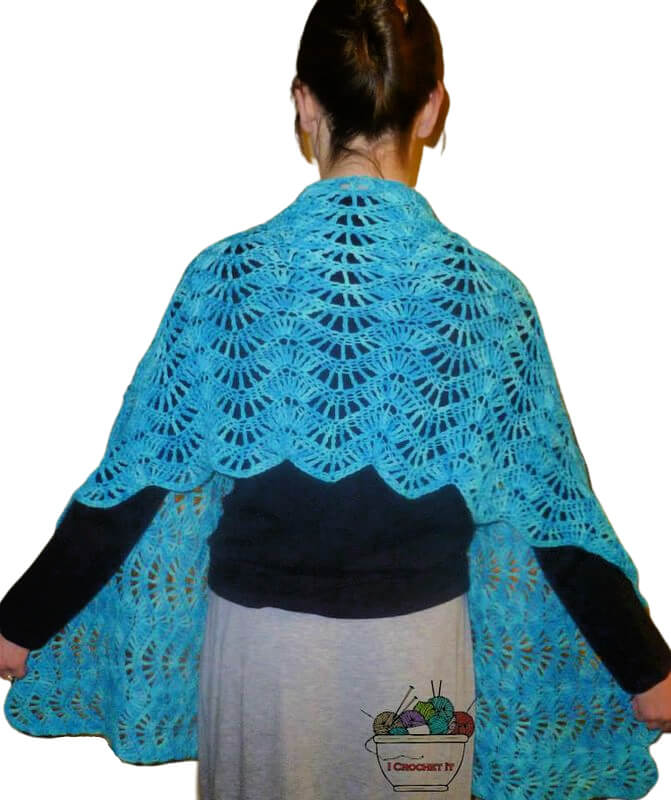 Back view from the hips up of a woman in a gray skirt and black top with a shawl wrapped around her shoulders. The shawl is made of a feather and fan crochet ripple stitch in medium blue yarn.