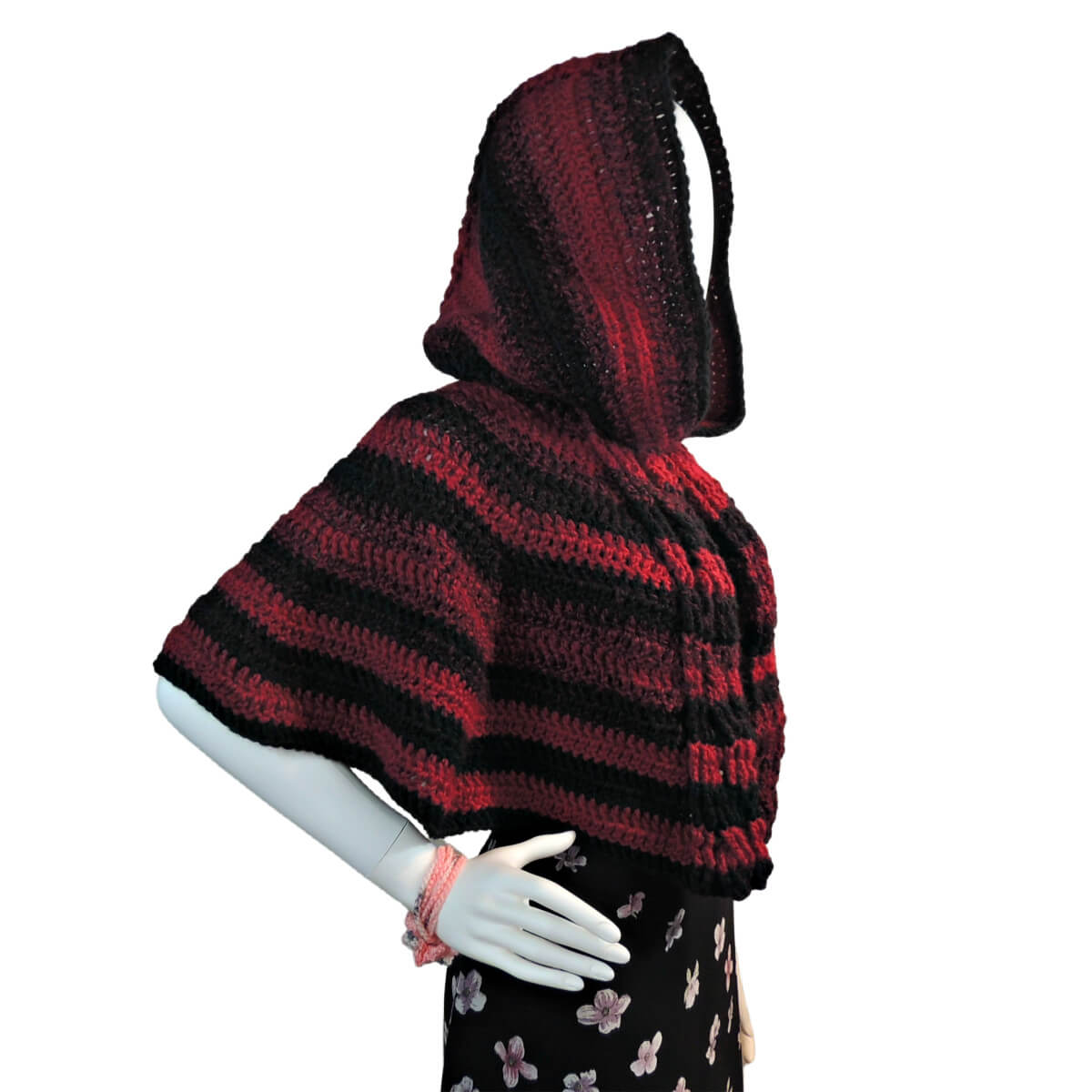 Side view of a white mannequin in a black skirt and a hooded waist length crocheted cape. The cape has cables down the front opening and horizontal stripes of black and red.