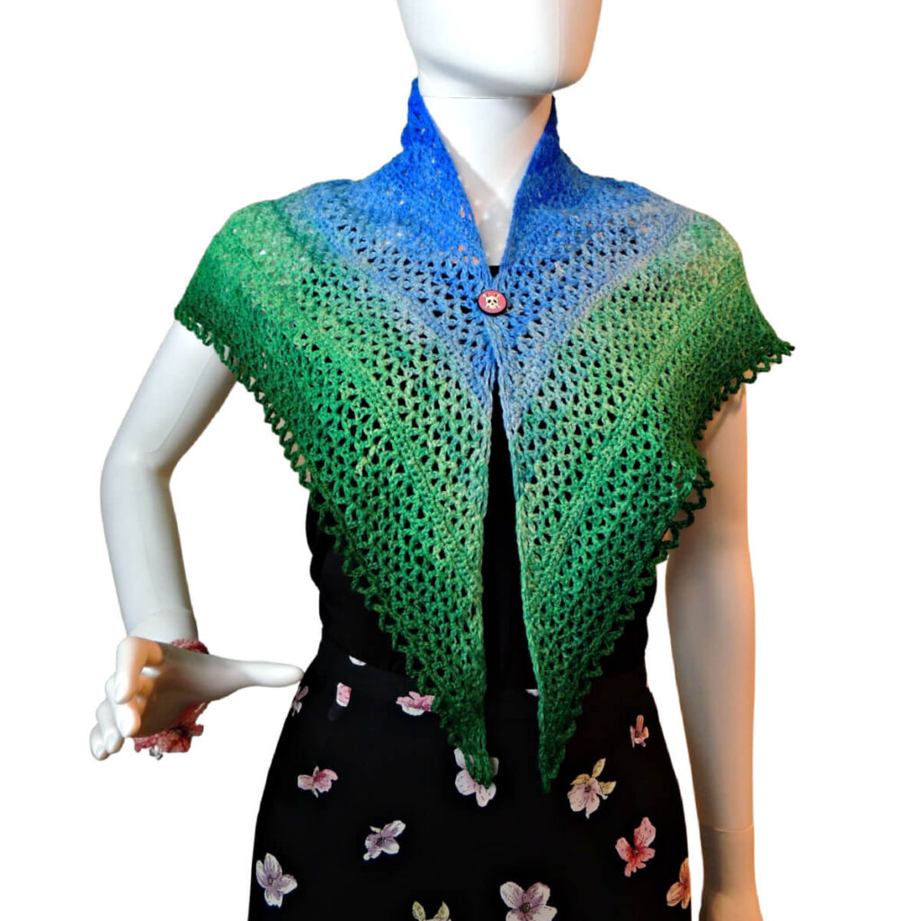 Waist up of a mannequin in a black tank top and skirt with a triangular crochet lace shawl with a scalloped chain edge draped over its shoulders and held together just under the neck with a small shawl button. The shawl changes color from the top center out from dark blue to light blue to light green to dark green.