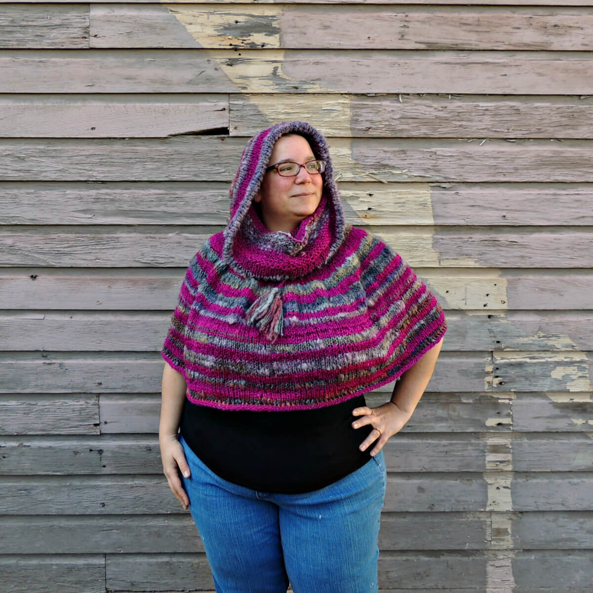 Woman standing in front of a wall with gray wood siding. She wears jeans, a black top and a tube shaped poncho with a drawstring about 1/3 down to create a hood above the drawstring and an elbow-length poncho below. The poncho is knit in yarn that that stripes between a thick and thin fuchsia and a fuzzy gray. The hood is pulled up on her head, and one hand is on her hip.