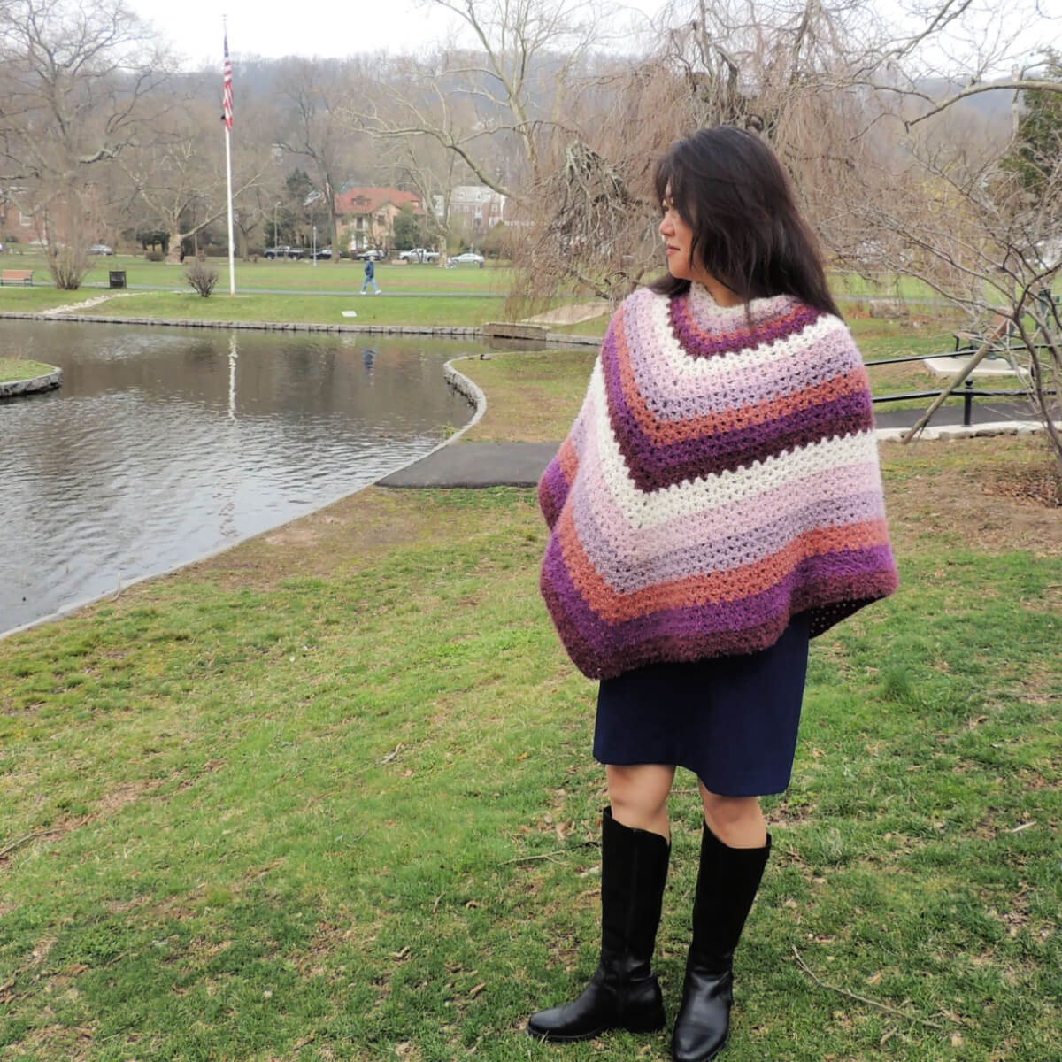 A woman in a park standing next to a small lake. She's wearing a black knee-length skirt, tall black boots, and a classic 2 point poncho crocheted in fuzzy yarn in stripes from the neck down of cream, lilac, lavender, light rust, purple, and burgundy, repeated so each color is seen 3 times. Her shirt can not be seen beneath the poncho.
