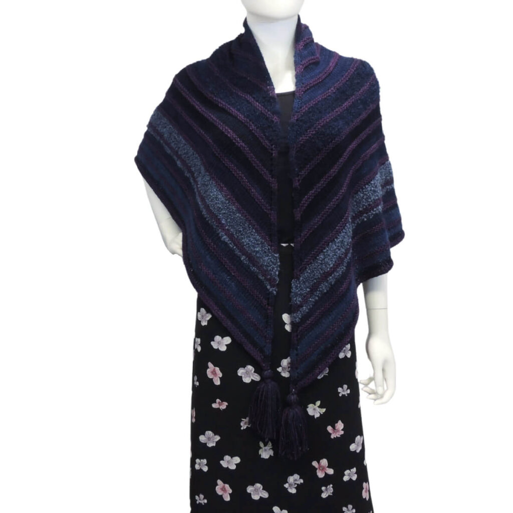 Front view of a white mannequin wearing a black skirt, black tank top, and a large triangular shawl in shades of blue with small raised stripes of purple every few inches, oversized tassels hang off the 3 points. The triangle shawl is a right angle with 2 equal sides, the center of the long side is at the back of the neck and wraps to the front where the 2 end points hang down.